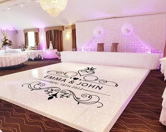 May include: A white dance floor with black lettering that reads "Emma & John, Nov. 18th 2022" with a decorative design. The dance floor is in a wedding reception hall with white tables and chairs.