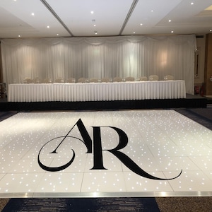 May include: An event venue with a white dance floor, illuminated with embedded lights, and a large black "AR" monogram. A long white table with chairs is set against a white draped backdrop. The room is well-lit, suitable for a wedding or formal occasion.