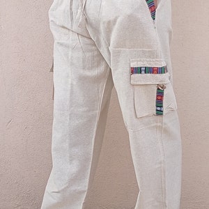 May include: Beige linen pants with a drawstring waist and colorful patterned trim on the pockets. The pants have a relaxed fit and are perfect for casual wear.