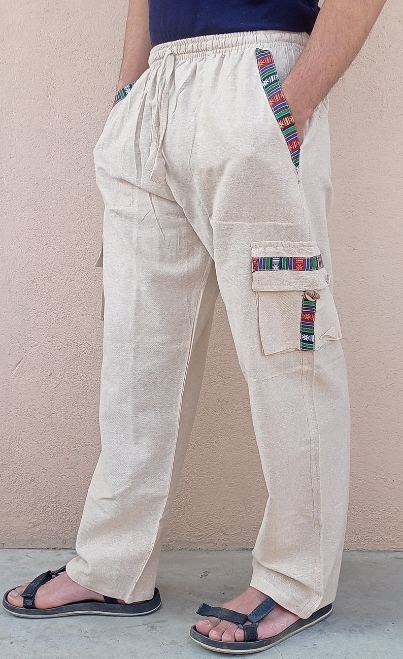 May include: Beige linen pants with a drawstring waist and colorful patterned trim on the pockets. The pants have a relaxed fit and are perfect for casual wear.