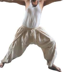May include: A person wearing a white tank top and beige loose-fitting pants, standing in a yoga pose with arms outstretched.