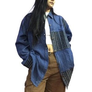 May include: A blue denim shirt with a patterned fabric panel on the front and cuffs. The shirt is unbuttoned and has a white undershirt visible. The person is wearing brown cargo trousers.