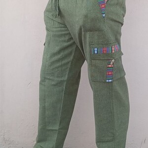 May include: Green cargo pants with a drawstring waist and colorful patterned trim. The pants have a large cargo pocket on the right leg with a flap and a decorative trim.