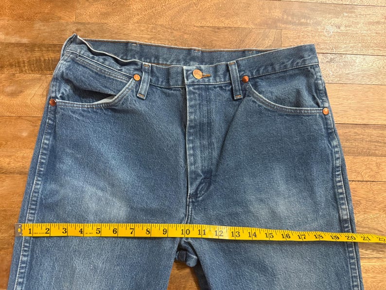 May include: Blue denim jeans with a straight leg cut. The jeans have a classic five-pocket design, copper rivets, and a button closure. A yellow measuring tape shows a length of approximately 20 inches.