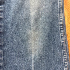 May include: Close-up of blue denim fabric. The denim has a faded appearance with lighter areas and vertical lines. The stitching is visible along the right side of the fabric.