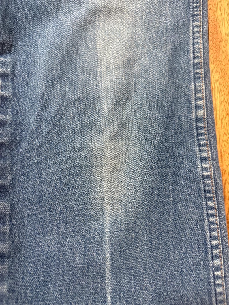 May include: Close-up of blue denim fabric. The denim has a faded appearance with lighter areas and vertical lines. The stitching is visible along the right side of the fabric.
