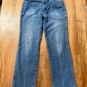 May include: Blue denim jeans with a classic five-pocket design. The jeans have a faded wash and a straight leg cut. The stitching is visible in a contrasting color, and the jeans are laid flat on a wooden surface.