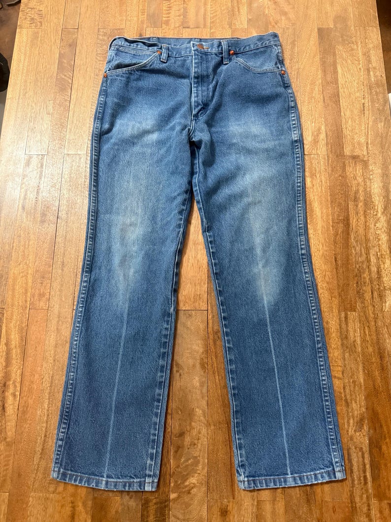 May include: Blue denim jeans with a classic five-pocket design. The jeans have a faded wash and a straight leg cut. The stitching is visible in a contrasting color, and the jeans are laid flat on a wooden surface.