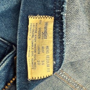 Pu&ograve; includere: Primo piano di jeans blu con un'etichetta del marchio Wrangler. L'etichetta indica una misura della vita di 86 cm e una lunghezza di 76 cm. I jeans sono etichettati come "Mens Regular Fit".