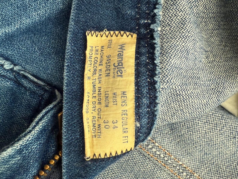 Pu&ograve; includere: Primo piano di jeans blu con un'etichetta del marchio Wrangler. L'etichetta indica una misura della vita di 86 cm e una lunghezza di 76 cm. I jeans sono etichettati come "Mens Regular Fit".