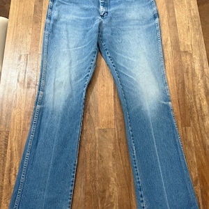 May include: Light blue denim jeans with a classic five-pocket design and a flared leg. The jeans have a button and zipper closure and are made of a durable denim material. The jeans are laid flat on a wooden surface.