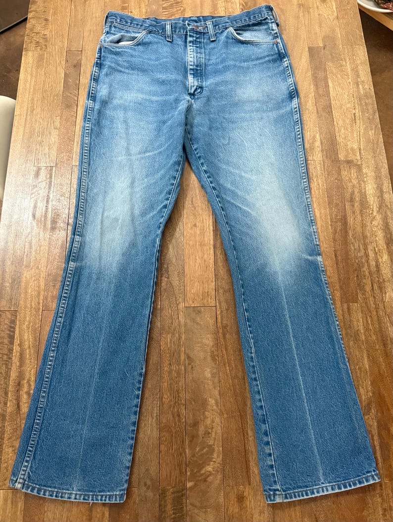 May include: Light blue denim jeans with a classic five-pocket design and a flared leg. The jeans have a button and zipper closure and are made of a durable denim material. The jeans are laid flat on a wooden surface.