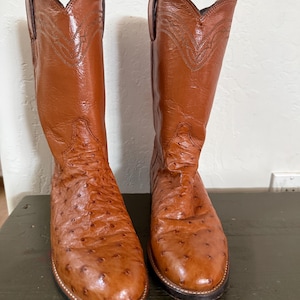 May include: A pair of brown leather cowboy boots. The boots have a textured surface and decorative stitching on the upper shaft. The soles are black with a light-colored stitch around the edge. The boots are displayed on a dark green surface.
