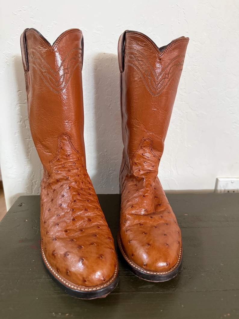 May include: A pair of brown leather cowboy boots. The boots have a textured surface and decorative stitching on the upper shaft. The soles are black with a light-colored stitch around the edge. The boots are displayed on a dark green surface.