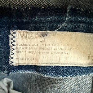 May include: Close-up of a denim garment with a white label stitched on. The label reads "Wrangler" and includes washing instructions: "Machine wash with like colors... tumble dry, remove promptly." The label also states "MADE IN USA."