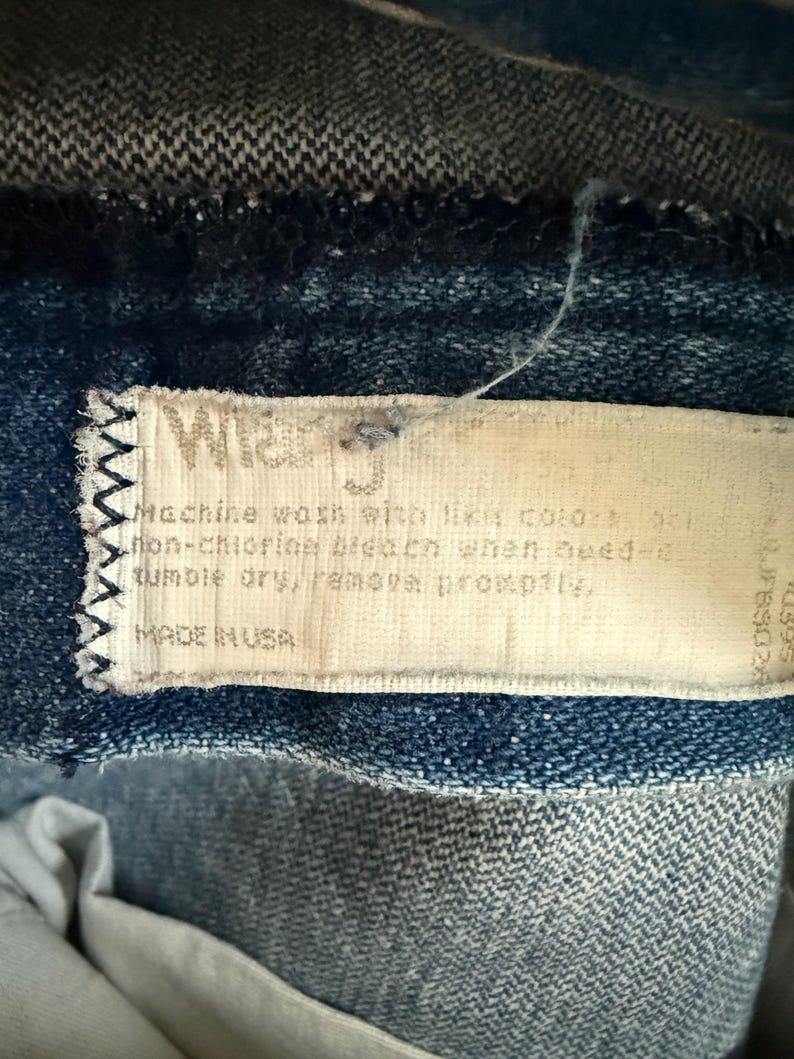 May include: Close-up of a denim garment with a white label stitched on. The label reads "Wrangler" and includes washing instructions: "Machine wash with like colors... tumble dry, remove promptly." The label also states "MADE IN USA."