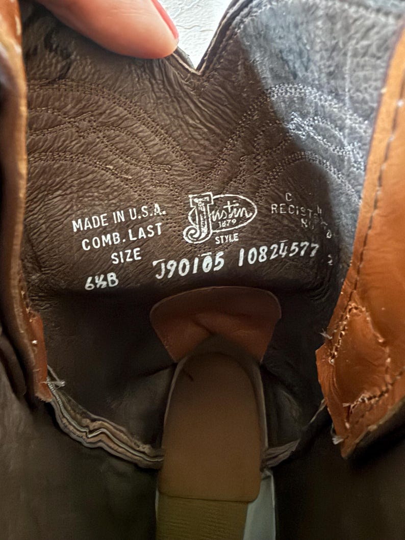 May include: Close-up of the inside of a brown leather cowboy boot. The boot's interior displays the brand name "Justin" and the text "Made in U.S.A.". The size is 6 1/2 B. The boot has a brown leather insole and a tan leather exterior.