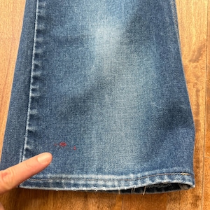 Pu&ograve; includere: Primo piano di jeans di denim blu con una leggera sbiaditura. I jeans hanno un classico design a cinque tasche e una gamba dritta. Un dito indica una piccola macchia rossa sul denim. I jeans sono su una superficie di legno.