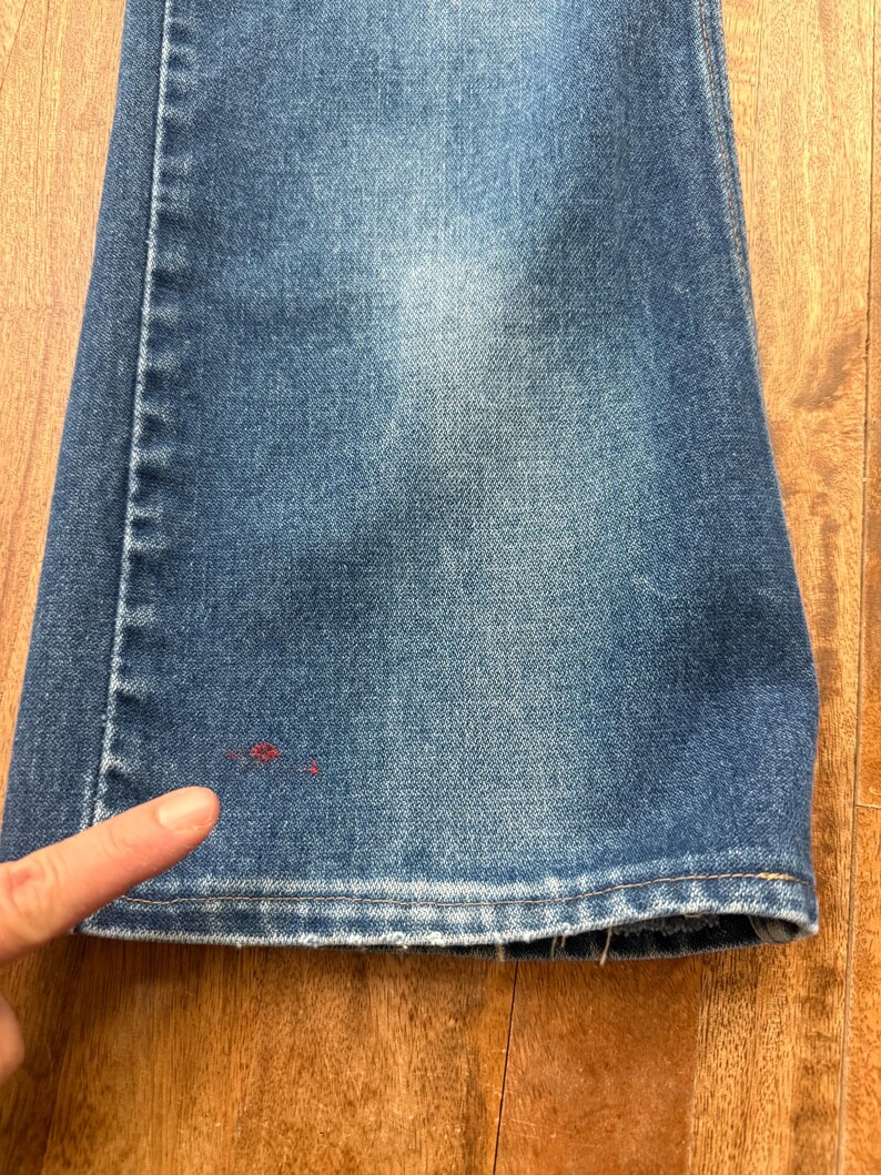 Pu&ograve; includere: Primo piano di jeans di denim blu con una leggera sbiaditura. I jeans hanno un classico design a cinque tasche e una gamba dritta. Un dito indica una piccola macchia rossa sul denim. I jeans sono su una superficie di legno.