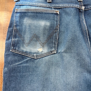 Pu&ograve; includere: Primo piano del retro di un paio di jeans blu. I jeans hanno un aspetto sbiadito con una tasca usurata con il logo Wrangler. Le cuciture sono visibili e il denim mostra segni di usura.