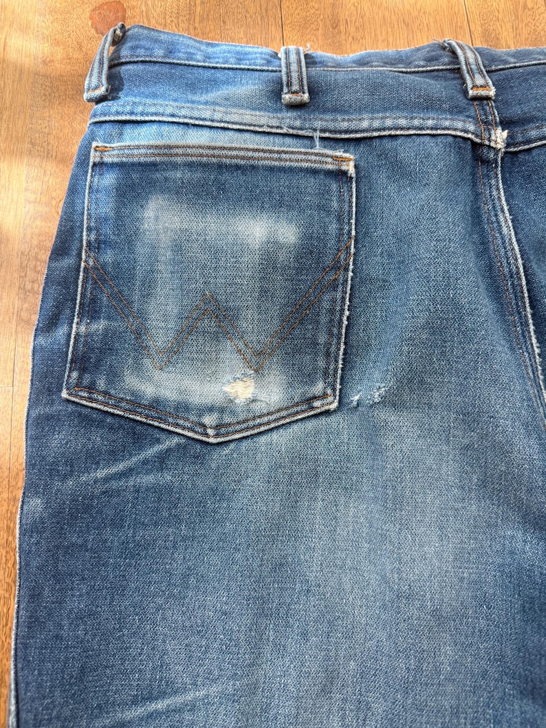 Pu&ograve; includere: Primo piano del retro di un paio di jeans blu. I jeans hanno un aspetto sbiadito con una tasca usurata con il logo Wrangler. Le cuciture sono visibili e il denim mostra segni di usura.