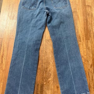 May include: Light blue denim jeans with two back pockets and a brown leather patch. The jeans have a classic five-pocket design and a straight leg cut. The Wrangler brand is visible on the leather patch.