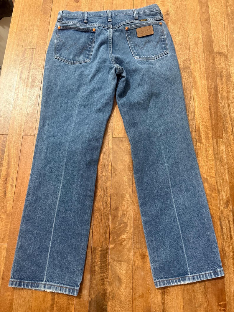 May include: Light blue denim jeans with two back pockets and a brown leather patch. The jeans have a classic five-pocket design and a straight leg cut. The Wrangler brand is visible on the leather patch.