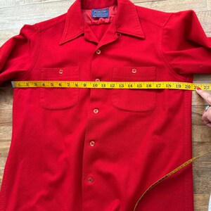 以下が含まれることがあります： 鮮やかな赤色のボタンダウンシャツで、胸ポケットが2つ付いています。 シャツは黄色のメジャーで測定されており、長さは約56cmです。 シャツには襟があり、前面にボタンが付いています。
