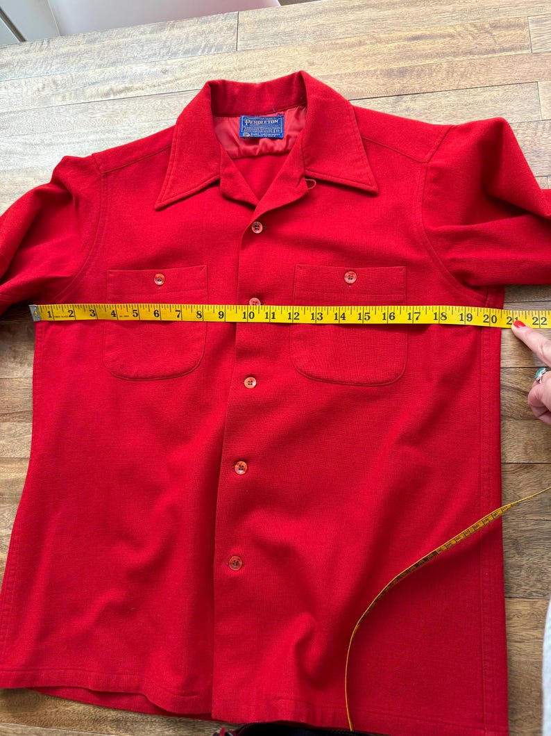 以下が含まれることがあります： 鮮やかな赤色のボタンダウンシャツで、胸ポケットが2つ付いています。 シャツは黄色のメジャーで測定されており、長さは約56cmです。 シャツには襟があり、前面にボタンが付いています。