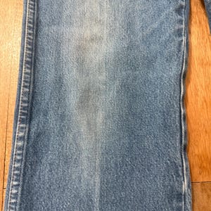 May include: Close-up of light blue denim jeans. The jeans show signs of wear and fading, with a lighter patch on the front. The stitching is visible along the seams. The jeans are on a wooden surface.