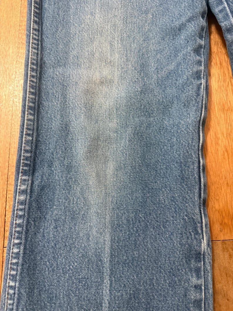 May include: Close-up of light blue denim jeans. The jeans show signs of wear and fading, with a lighter patch on the front. The stitching is visible along the seams. The jeans are on a wooden surface.