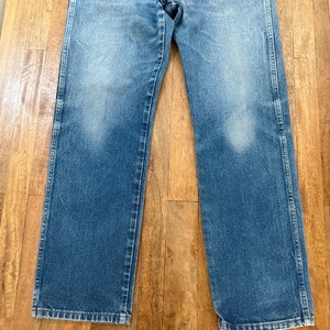 K&ouml;nnte beinhalten: Verblasste blaue Jeans mit geradem Bein. Die Jeans weisen Abnutzungserscheinungen mit helleren Flecken auf der Vorderseite der Beine auf. Die Jeans liegen flach auf einer Holzoberfl&auml;che.