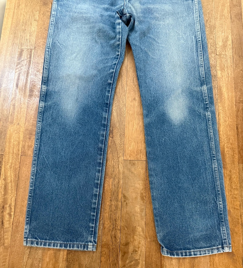K&ouml;nnte beinhalten: Verblasste blaue Jeans mit geradem Bein. Die Jeans weisen Abnutzungserscheinungen mit helleren Flecken auf der Vorderseite der Beine auf. Die Jeans liegen flach auf einer Holzoberfl&auml;che.