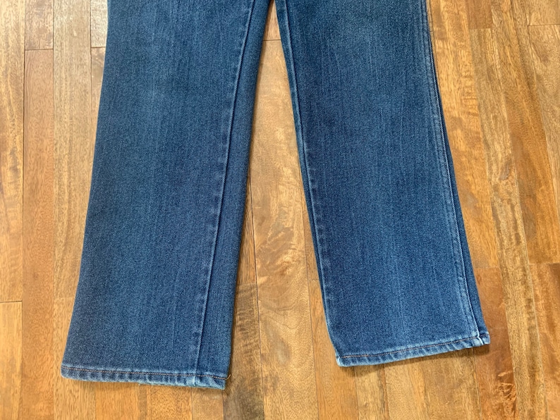 May include: A pair of blue denim jeans with a wide leg opening. The jeans are laying flat on a wooden surface.
