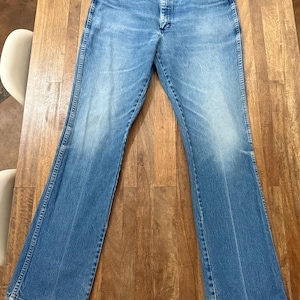 May include: Light blue denim jeans with a classic five-pocket design. The jeans have a faded wash and a slight flare at the hem. The jeans are laid flat on a wooden surface.
