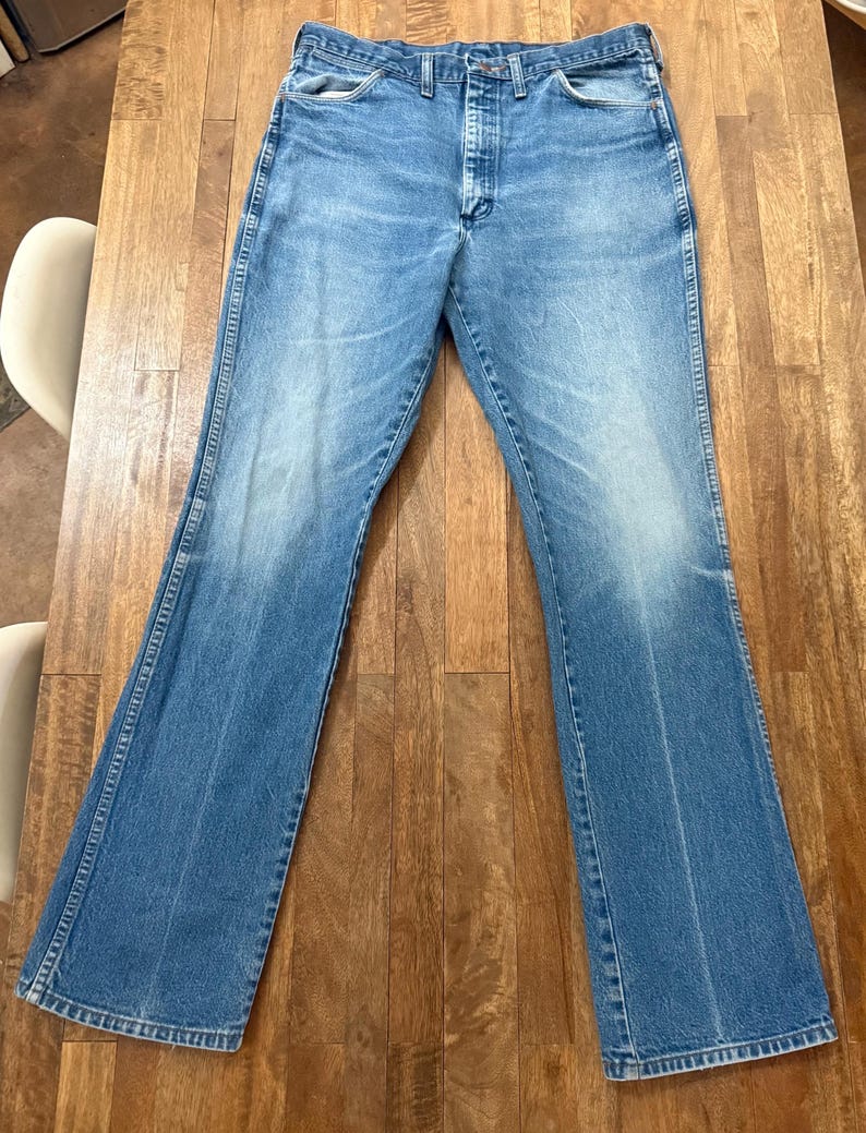 May include: Light blue denim jeans with a classic five-pocket design. The jeans have a faded wash and a slight flare at the hem. The jeans are laid flat on a wooden surface.