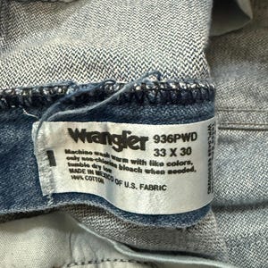 May include: Close-up of a blue denim Wrangler jeans label. The label displays the brand name, size 33 x 30, and washing instructions. The jeans are made of 100% cotton and manufactured in Mexico of U.S. fabric.