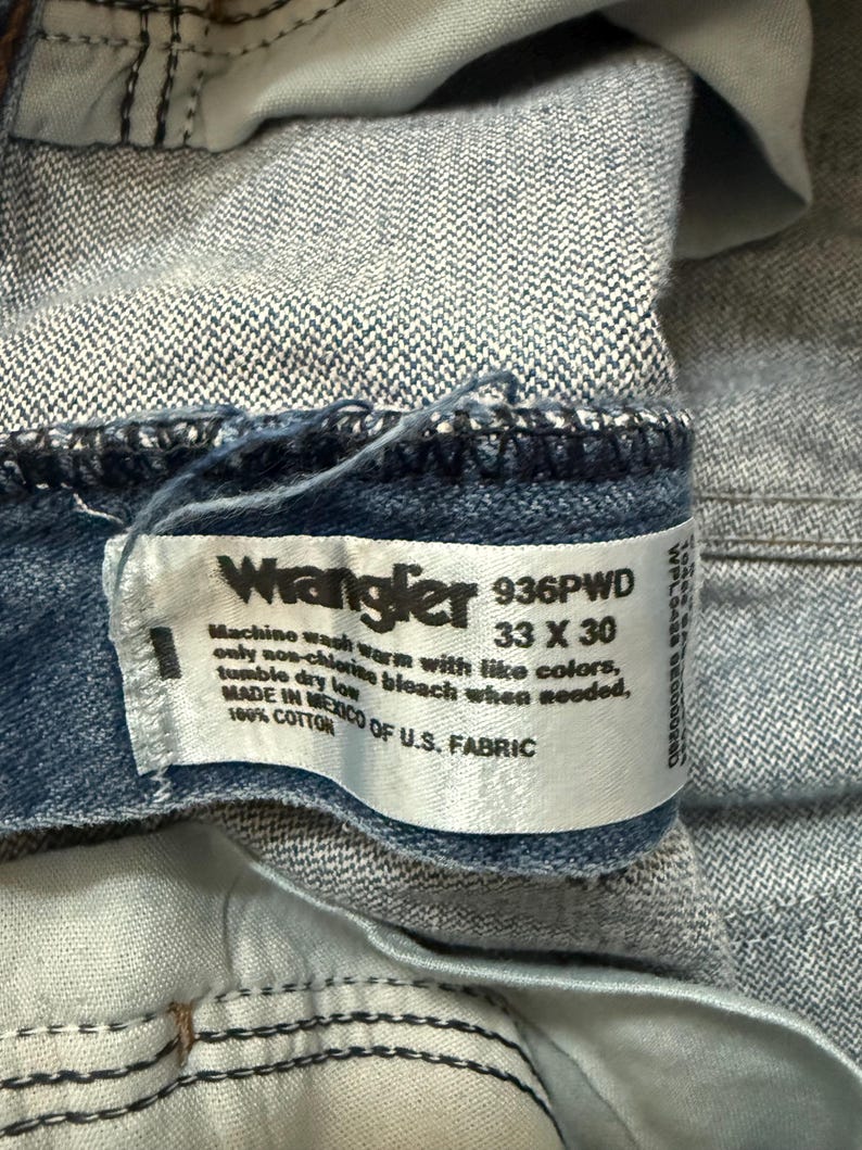 May include: Close-up of a blue denim Wrangler jeans label. The label displays the brand name, size 33 x 30, and washing instructions. The jeans are made of 100% cotton and manufactured in Mexico of U.S. fabric.