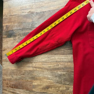 以下が含まれることがあります： ポケット付きの赤い長袖シャツを黄色のメジャーで測定しています。袖の長さは約58cmです。シャツは木製の表面にあります。