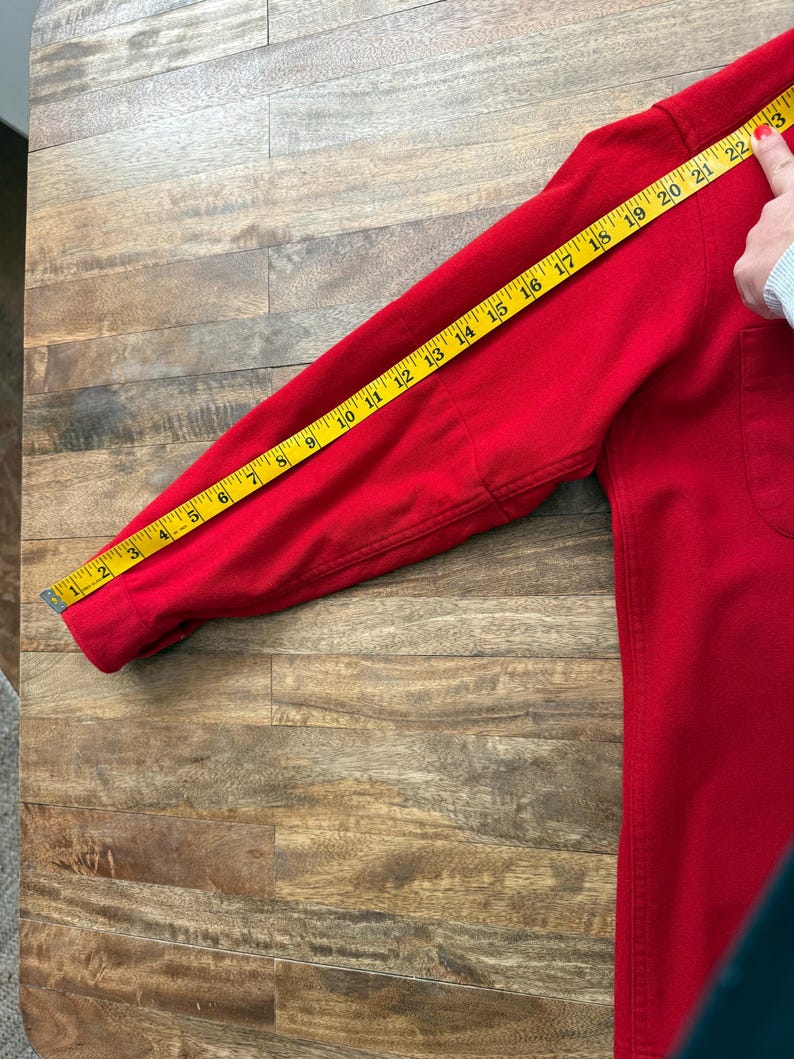 以下が含まれることがあります： ポケット付きの赤い長袖シャツを黄色のメジャーで測定しています。袖の長さは約58cmです。シャツは木製の表面にあります。