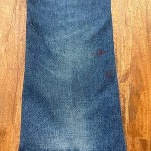 Pu&ograve; includere: Primo piano di jeans di denim blu con una leggera svasatura sul fondo. I jeans hanno un aspetto sbiadito con alcune macchie pi&ugrave; scure vicino al lato destro. Il tessuto mostra una classica trama denim.