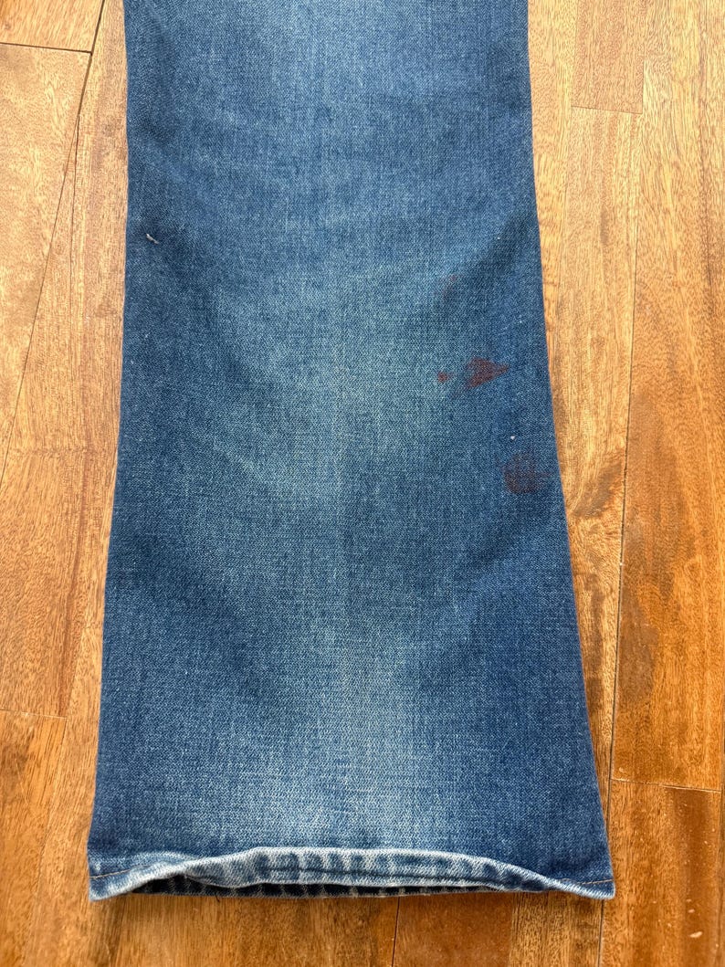 Pu&ograve; includere: Primo piano di jeans di denim blu con una leggera svasatura sul fondo. I jeans hanno un aspetto sbiadito con alcune macchie pi&ugrave; scure vicino al lato destro. Il tessuto mostra una classica trama denim.