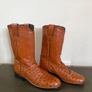 May include: A pair of brown leather cowboy boots. The boots have a textured pattern on the foot and a smooth finish on the upper shaft. The boots have a classic western design with a pointed toe and a stacked heel.