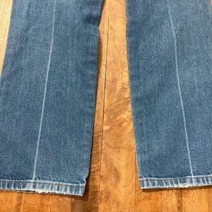 May include: Close-up of a pair of blue denim jeans. The jeans have a light blue wash with a vertical seam down the center of each leg. The hem is frayed. The jeans are on a wooden surface.