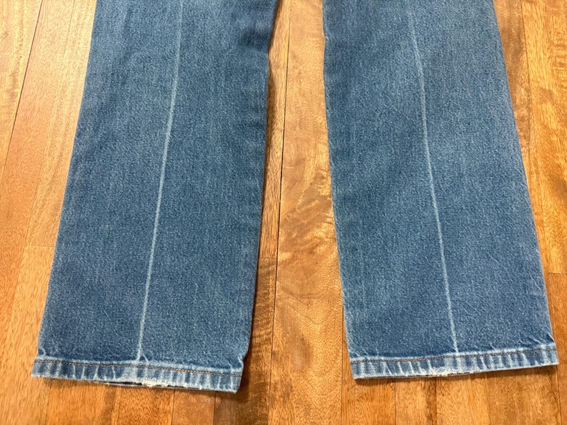 May include: Close-up of a pair of blue denim jeans. The jeans have a light blue wash with a vertical seam down the center of each leg. The hem is frayed. The jeans are on a wooden surface.