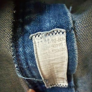 May include: Close-up of a blue denim garment with a sewn-in tag. The tag is rectangular, with text including numbers and letters. The denim fabric is a dark blue wash, and the tag is a light beige color. The tag is sewn into the garment with a zigzag stitch.