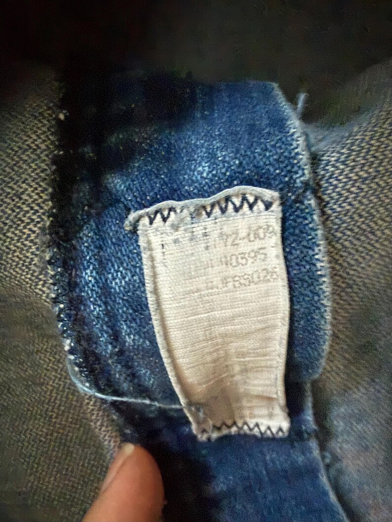 May include: Close-up of a blue denim garment with a sewn-in tag. The tag is rectangular, with text including numbers and letters. The denim fabric is a dark blue wash, and the tag is a light beige color. The tag is sewn into the garment with a zigzag stitch.
