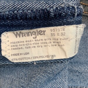 May include: A white tag with black text attached to a pair of blue denim jeans. The tag reads "Wrangler 937STR 35 X 32", "Machine wash warm with like colors, only non-chlorine bleach when needed, tumble dry low, low iron", "MADE IN USA", "67% COTTON/33% POLYESTER".