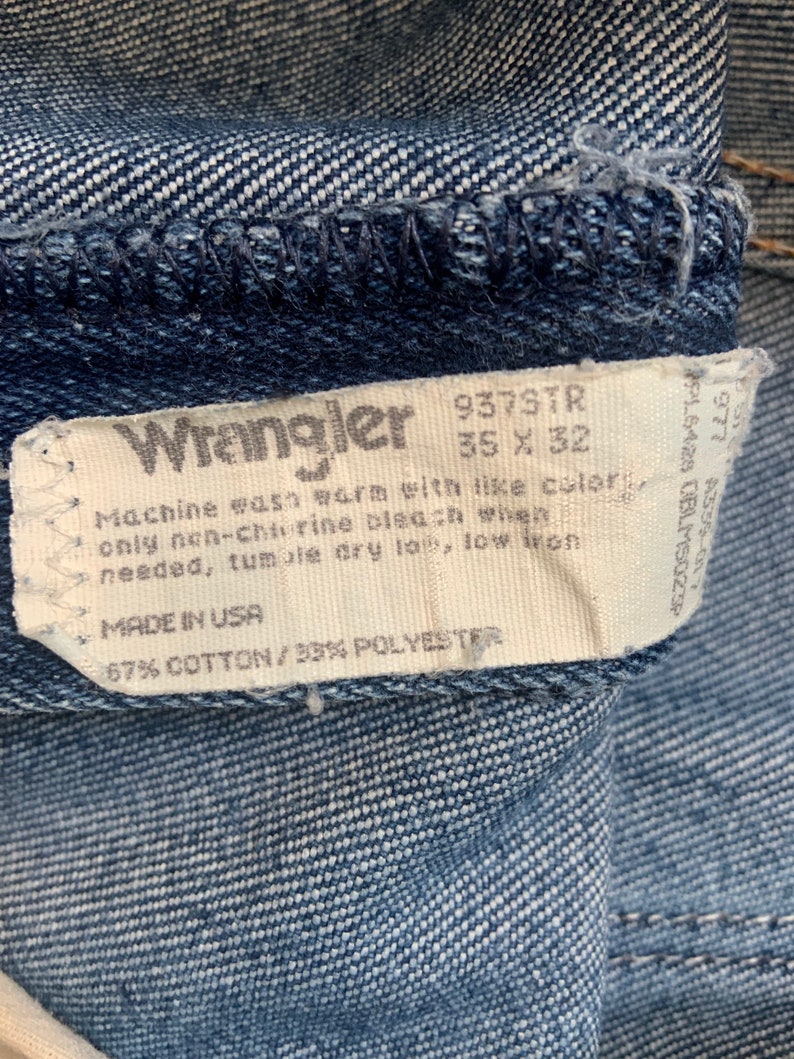 May include: A white tag with black text attached to a pair of blue denim jeans. The tag reads "Wrangler 937STR 35 X 32", "Machine wash warm with like colors, only non-chlorine bleach when needed, tumble dry low, low iron", "MADE IN USA", "67% COTTON/33% POLYESTER".