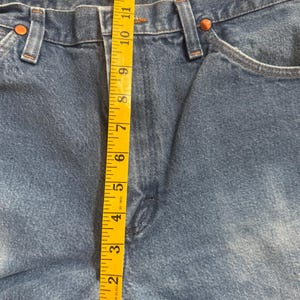 May include: Close-up of blue denim jeans with a yellow measuring tape. The tape shows a measurement of approximately 13 inches. The jeans have copper rivets and a classic five-pocket design.